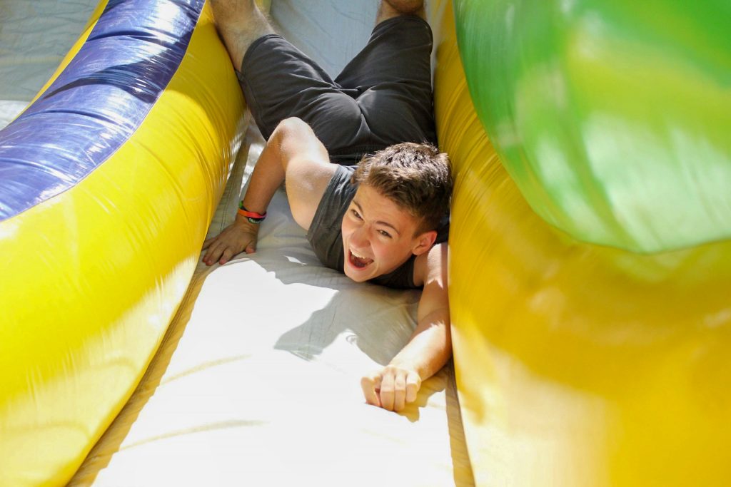 A camper plays on an inflatable obstacle course. He is smiling and falling down the inflatable toy.